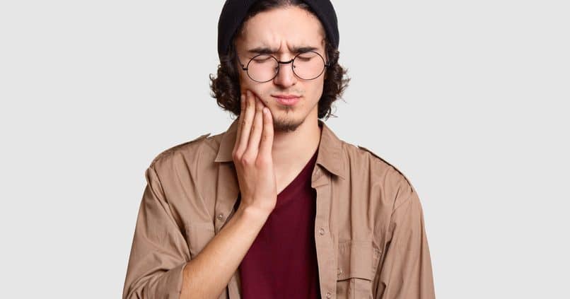 photo-of-stressful-youngster-with-small-beard-keeps-hand-on-cheek-suffers-from-toothache-keeps-eyes-shut-dressed-in-stylish-clothes-big-round-spectacles-models-against-white-studio-wall